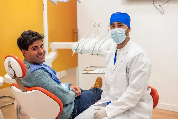 Cerrahi maske takan genç erkek dişçi, diş sağlığı ve diş sağlığını vurgulayan modern bir diş sağlığı kliniğindeki bir dişçi koltuğunda rahatça otururken, güvenli bir şekilde poz veriyor.