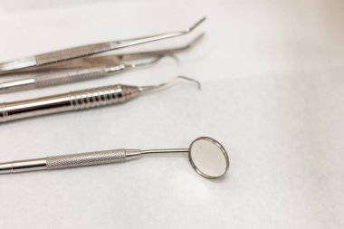 Ağız aynası, problar ve cımbızlar da dahil olmak üzere çeşitli metal diş aletleri beyaz bir yüzeyde, diş sağlığı ve ağız sağlığı hizmetlerini temsil eden bir diş kliniğinde kullanılmaya hazırdır.