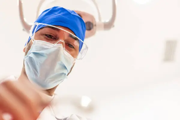 Cerrahi maske, gözlük ve mavi cerrahi başlık takan POV Dişçi, modern bir klinik ortamındaki hastalara kaliteli oral sağlık hizmeti sağlamak için hassas bir diş prosedürü uyguluyor.