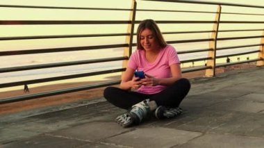 Kumsalda bacak bacak bacak üstüne atmış genç bir kadın, altın gün batımında dahili paten giyerken akıllı telefon kullanıyor, okyanus arkaplanının keyfini çıkarıyor.