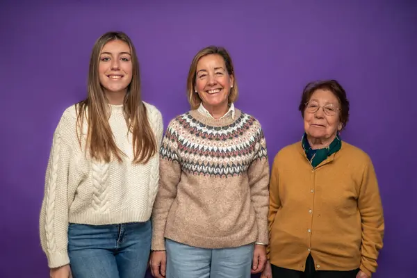 Büyükanne, anne ve kız birlikte poz veriyorlar. Aile birliği ve kadın gücünü gösteriyorlar. Mor arka planda kışlık kıyafetler giyiyorlar.