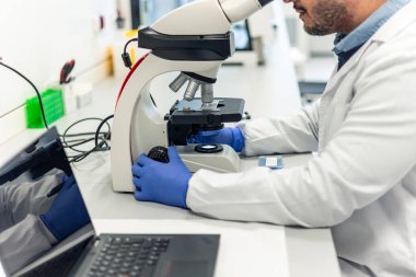 Laboratuar önlüklü ve mavi eldivenli bilim adamı mikroskop altında örnekleri incelerken modern bir laboratuvarda veri analizi için yakınlarda bir dizüstü bilgisayar bulunduruyor.