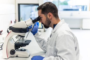 Laboratuvar önlüğü ve eldiven giyen genç bir bilim adamı mikroskoptan bakıp laboratuvarda mikro pipet kullanarak bilimsel araştırma ve analiz yapıyor.
