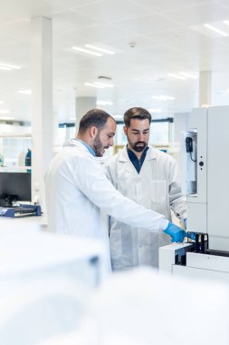 Laboratuvar önlüklü iki erkek bilim adamı yenilikçi araştırmalar üzerinde çalışıyor ve ileri laboratuvar ekipmanlarıyla çalışırken sonuçları tartışıyorlar.