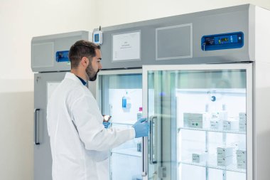 Laboratuvar önlüğü ve eldiven giyen bilim adamı, ileri düzey araştırma ekipmanları ve teknoloji ile çevrili modern bir laboratuvarda ilaç kutularıyla dolu bir buzdolabı açıyor.