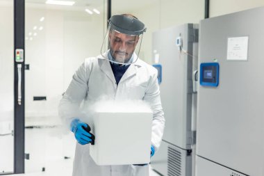 Koruyucu laboratuvar önlüğü, eldiven ve yüz kalkanı giyen bilim adamı kriyojenik dondurucularla dolu bir laboratuarda dumanı tüten kuru bir buz kutusu taşıyor.