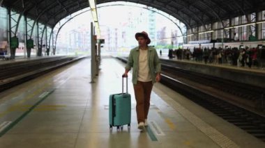 Platform boyunca yürüyen şapkalı ve mavi valizli bir gezgin, trenlerinin gelmesini beklerken kameraya doğru yürüyor, 4K video.