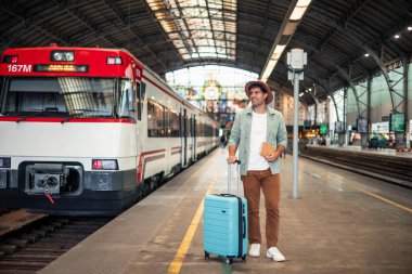 Young latin tourist wearing a hat waits at the train station, holding a suitcase and passport while gazing into the distance, anticipating the next adventure on his journey clipart