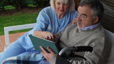 Şefkatli sağlık uzmanı, yaşlı hastaya destek olurken dijital tablet kullanıyor, anlamlı etkileşim ve bağlantı sırasında güneşli sundurmada yan yana oturuyor.