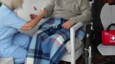 Profesyonel bakıcı, tekerlekli sandalyedeki yaşlı adamın elini dikkatlice bandajlıyor. Ev sağlığı tedavisi sırasında şefkatli tıbbi destek sağlıyor.