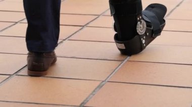 Ortopedik yürüyüş botu giyen hasta fayanslı yüzeyde yavaşça hareket ediyor. Alt bacak yaralanmasından sonra dikkatli bir rehabilitasyon ve iyileşme gösteriyor.