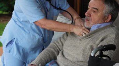 Uzman hemşire steteskop kullanarak yaşlı hastaya göz kulak oluyor ve evde yapılan tıbbi muayene sırasında şefkatli bir ihtiyari sağlık desteği sağlıyor.