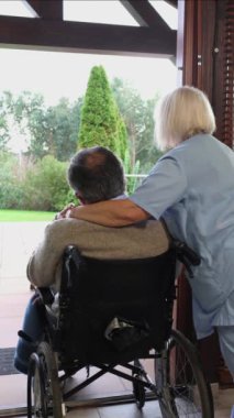 Tekerlekli sandalyedeki yaşlı hastayı kucaklarken duygusal destek sunan şefkatli hemşire, huzur dolu bahçeye empati ve özenle bakıyor.