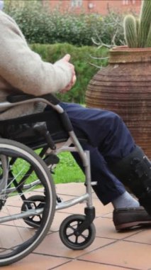 Dikey Yaşlılar bacak yaralanmasından kurtuluyor ortopedik yürüyüş botu giyiyor, huzurlu bahçe ortamında tekerlekli sandalyede otururken rehabilitasyon egzersizleri yapıyor.
