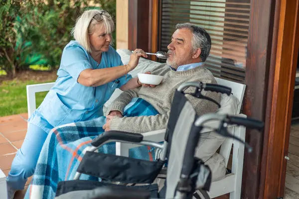 Uzman bir bakıcı, tekerlekli sandalyedeki kıdemli bir adama yardım ediyor, ona rahat bir verandada yemek veriyor, ev sağlığı ve yaşlılara destek olmanın önemini vurguluyor.