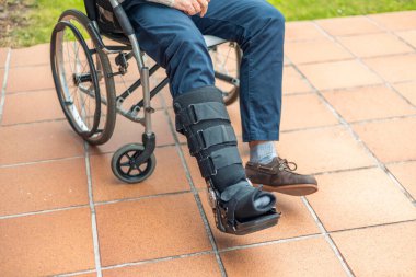 Bacak korsesi takan ve tekerlekli sandalyede oturan hasta bir kliniğin ya da hastanenin avlusunda dinleniyor. Yaraları iyileşiyor ve güneş ışığının keyfini çıkarıyor.
