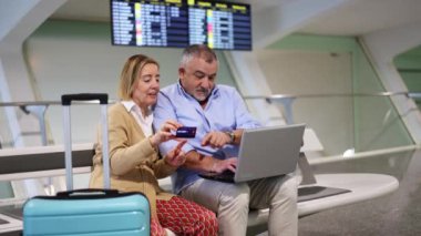 Üst düzey turist çift havaalanı terminalindeki bankta dizüstü bilgisayar ve kredi kartıyla oturuyor, arka planda uçuş bilgileri var.