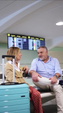 Dikey yaşlı turistler bir bankta oturuyor, sohbet ediyorlar ve sabırla uçuşlarını bekliyorlar, bavulları yanlarında ve arka planda uçuş bilgileri sergileniyor.