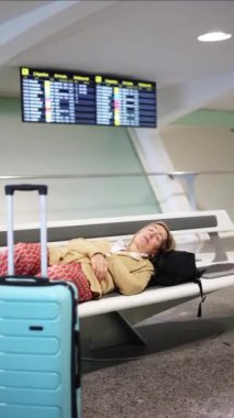 dikey kıdemli turist kadın havaalanı terminalinde dinleniyor, açık mavi tramvay valizi ve siyah sırt çantasının yanında uyuyor, kalkışı bekliyor.