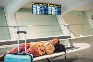 Havaalanı terminalinde, açık mavi tramvayı ve sırt çantasının yanındaki bankta uyuyan son sınıf öğrencisi kadın.