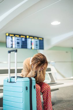 Bir havaalanı terminalinde bir bankta oturan dikey yaşlı kadın, gecikmiş bir uçuş yüzünden elleri sıkıntılı bir şekilde yüzünü kapatıyor, yanında açık mavi bir tramvay valiziyle