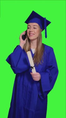Mavi önlük ve mezuniyet şapkası takan, elinde diploma olan, yeşil ekran karşısında dikilirken telefonla iletişim kuran akademik başarıyı neşeyle kutlayan yüksek lisans öğrencisi.