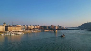 Panoramik hava aracı görüntüsü Budapeşte 'nin ikonik Zincir Köprüsü' nü yakalıyor, Tuna nehri boyunca süzülen manzaralı tekne kentsel ufuk çizgisi ve parlak mavi gökyüzü altında mimari simgeler