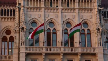 02-17-2025, Budapeşte. Macaristan ulusal bayrakları Budapeşte Parlamento Binası 'nın süslü Gotik cephesine karşı zarif bir şekilde dalgalanıyor. Gündüz vakti ulusal gurur ve yasama gücünü simgeliyor.