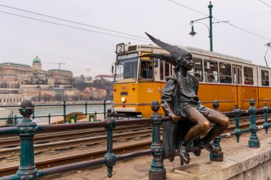 02-17-2025, Budapeşte. Küçük prensesin bronz heykeli tramvay raylarında oturmuş, Budapeşte, Macaristan 'daki Tuna Nehri ve Buda Kalesi' nin önünden geçen sarı tramvayı izliyor.