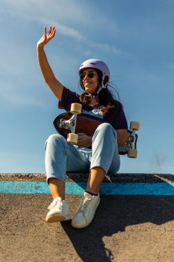 Kask takan ve güneş gözlüğü takan genç Latin bir kadın. Pompalı bir pistte oturmuş elinde kaykayla güneşli bir günün tadını çıkarıyor. En sevdiği sporu yapıyor.