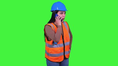Turuncu yelek ve mavi kasklı inşaat işçisi bir telefon görüşmesi yapıyor, gülümsüyor ve başka tarafa bakıyor, yeşil ekranda izole edilmiş.