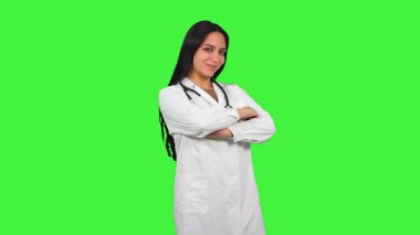 Beyaz laboratuvar önlüğü ve steteskop giyen, yeşil ekrana karşı kollarını kavuşturarak gülümseyen, profesyonel tıbbi uzmanlık sergileyen kendine güvenen Latin kadın doktor.