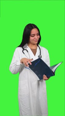 Beyaz laboratuvar önlüğü ve steteskop giyen dikey Latin kadın doktor yeşil ekran arka planına karşı tıbbi kayıtları okuyor, klinik uzmanlık ve odaklanmış tıbbi analiz gösteriyor