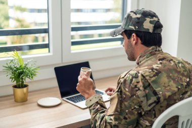 Kamuflaj üniformalı ve şapkalı bir Amerikan askeri masasında oturuyor, evde çalışıyor, kahve içiyor ve penceresinin yanında panjurları ve küçük bitkileri olan boş ekranlı dizüstü bilgisayar kullanıyor.
