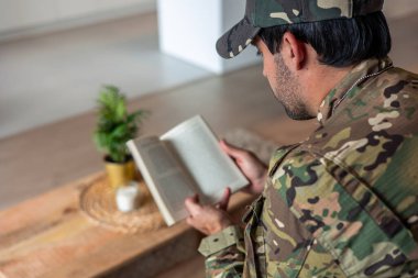 Kamuflaj üniformalı depresif bir gazi kitap okuyor, oturma odasında bir koltukta oturuyor, travma sonrası stres bozukluğundan muzdarip, rahatlamaya ve iyileşmeye çalışıyor.