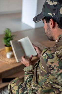 Kamuflaj üniforması ve şapka giyen genç bir asker evdeki koltukta otururken kitap okuyor, travma sonrası stres bozukluğundan muzdarip, iyileşmeye çalışıyor.