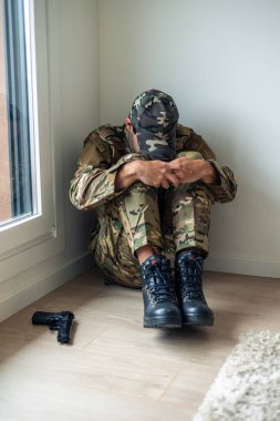 Kamuflaj üniformalı genç bir asker yerde oturuyor, dizlerinin arasında, yakın bir silahla, savaş sonrası travma sonrası stres bozukluğu yaşıyor.
