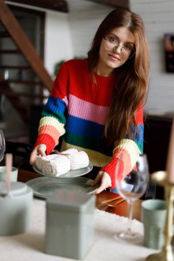 Kış süveteri giymiş gülümseyen bir kadın, davetlilerle yemek yemeden önce masayı hazırlar. Geleneksel aile tatili.