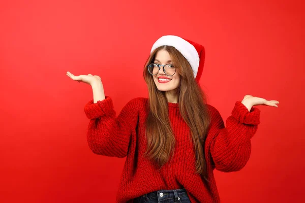 A beautiful girl makes a choice, shows her hands. Christmas gifts, sale, holiday offer. New Year\'s sweater and santa\'s hat on a red background.