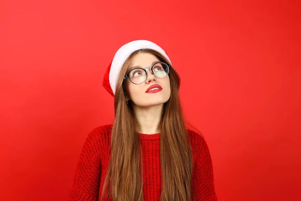 Portrait of an attractive girl in a santa hat and a Christmas sweater thinks about choosing a New Year's gift. Woman dreaming on a red background.