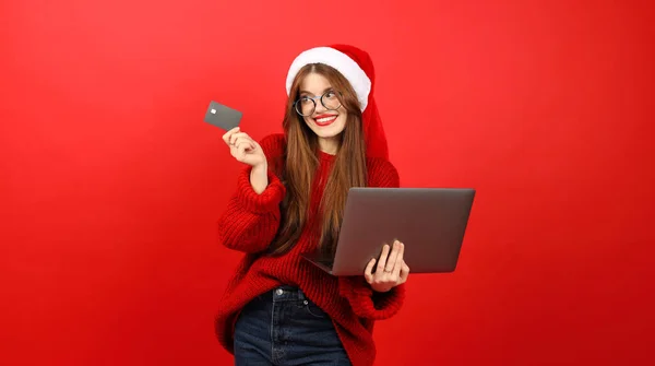 Happy consumer at christmas. Beautiful positive woman in santa hat and red sweater orders gifts on laptop.