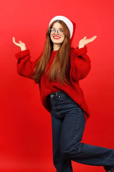Confused and surprised young woman in santa hat on red background. New Year\'s fuss.