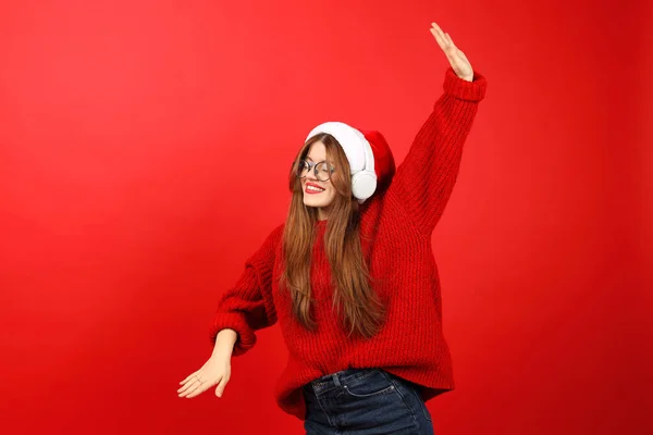 A beautiful young woman dances carelessly and enjoys Christmas music in headphones on a red background. Holiday dance playlist.