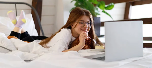 stock image Cute girl watching movie and eating pie, cake in bed, weekend vacation. Streaming services and series.