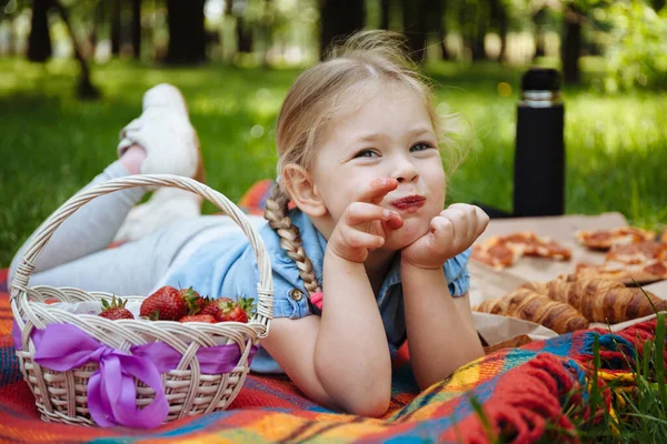 Cheerful little girl eating strawberries at picnic in park, sitting on green grass and smiling. Summer holidays and recreation.