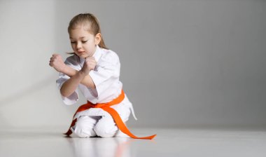 Küçük kız beyaz arka planda bir kimonoyla oturuyor, karate dersi..