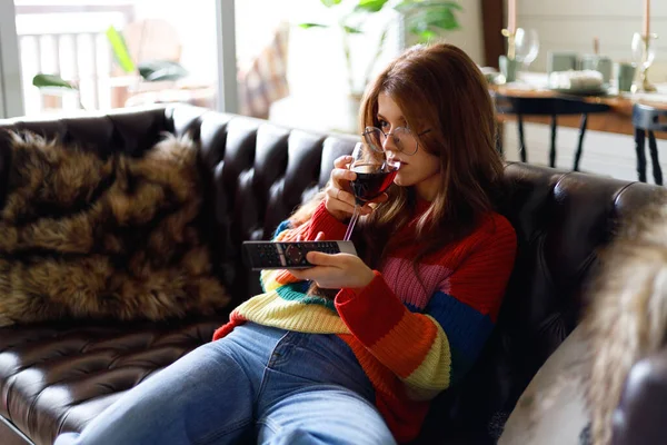 Attractive young girl is watching TV series with a glass of wine in a cozy home interior.