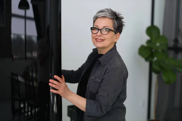 stock image Positive adult woman opens the door of a dark refrigerator, chooses food.