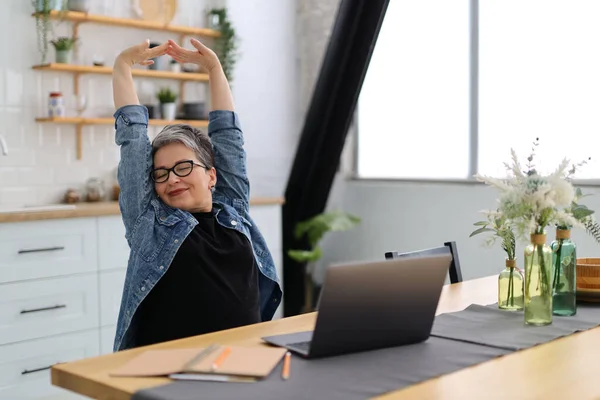 stock image A mature woman in glasses stretches at a table with a laptop.