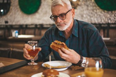 Mature man at the bar drinking beer and eating barbecue sandwich. Alcohol and hearty food, rest in a tavern.